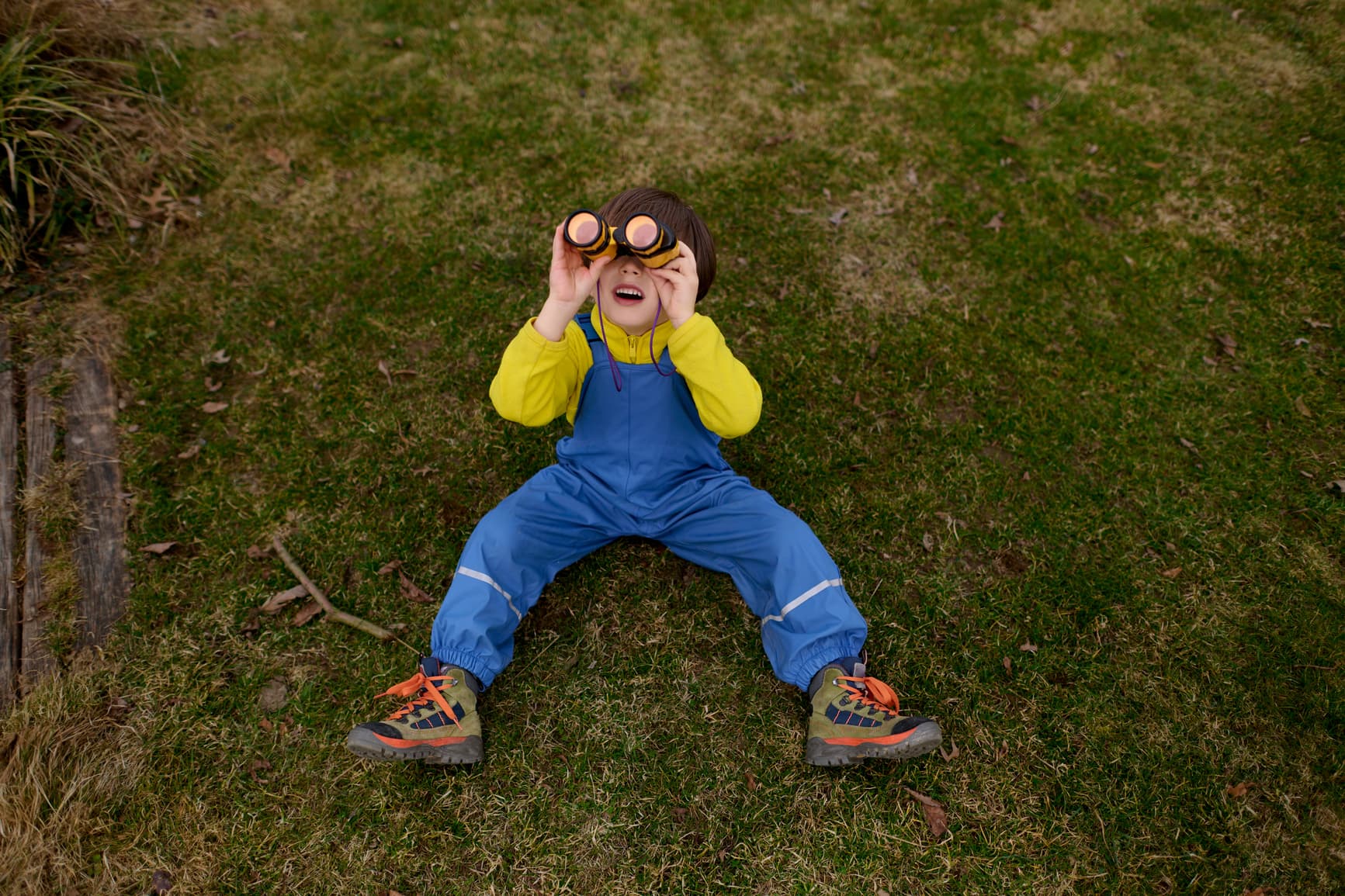 Cute young boy lying on green grass and looking up through binoculars, enjoying outdoor exploration and playful discovery in nature.